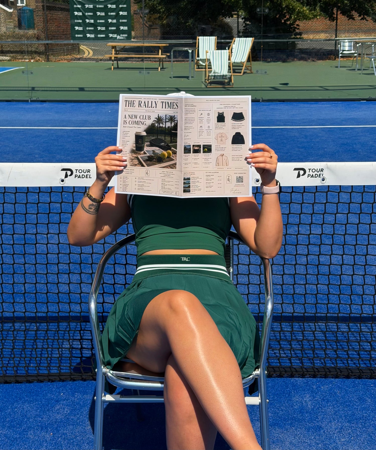 A woman sits on a chair on a blue tennis court, holding a newspaper in front of her face. She is wearing a dark green athletic dress with white trim.
